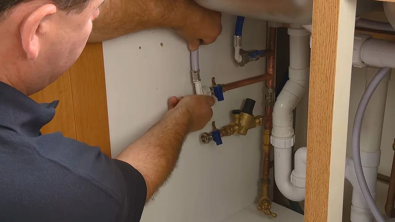 A man in a navy shirt reaching under a sink cabinet to adjust copper supply valves beside a white drain pipe